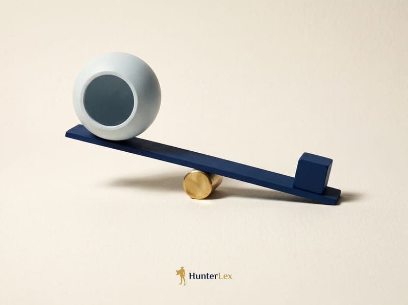 A navy lever balanced on a brass fulcrum, a large hollow pale sphere raised on one end and a small solid navy cube holding the other end down
